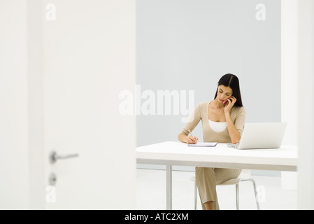 Geschäftsfrau sitzen am Schreibtisch, mit Handy und Laptop-Computer, auf dem Papier zu schreiben Stockfoto