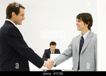 Zwei männliche Profis Händeschütteln, Lächeln einander, Kollege sitzt im Hintergrund Stockfoto