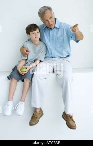 Mann, sitzend mit Arm um Enkels Schultern zeigend, beide wegschauen Stockfoto