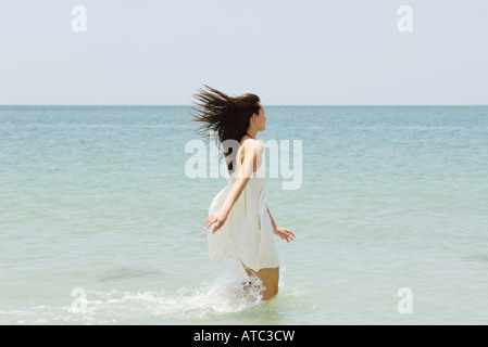 Junge Frau am Strand, Knie tief im Wasser, Rückansicht Stockfoto