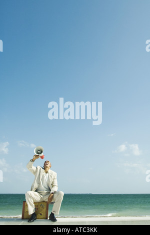 Mann sitzt auf Koffer am Strand, in Megaphon schreien Stockfoto