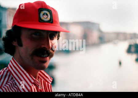 Mann in einem Sriped Hemd und Baseball Kappe sowohl rote lächelnd in die Kamera mit Canal in Venedig als Hintergrund Stockfoto