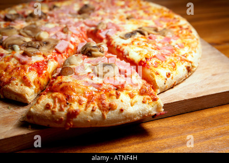 Eine große Käse-, Schinken- und Pilzpizza mit einer Scheibe, die fertig zum Essen ist. Pizza ist auf einem Holzbrett und wirkt sehr appetitlich und lecker. Stockfoto