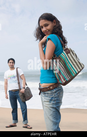 Porträt einer jungen Frau mit einer Handtasche mit einem jungen Mann stehend im Hintergrund am Strand Stockfoto