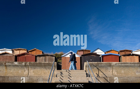 Frinton am Meer Strandhütten hinter der geschwungenen konkreten Seawall und ein einsamer Wanderer, vor blauem Himmel Stockfoto