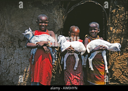 Drei 3 Mädchen hält Baby Ziegen außerhalb ihrer traditionellen Schlamm und Flechtwerk-Hütte in der Nähe von Masai Mara National Reserve Kenia in Ostafrika Stockfoto