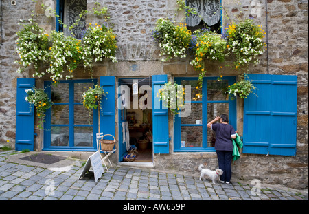 Frau guckend durch das Fenster eines hübschen, blau verschweißten Ladens mit hängenden Körben und ihrem kleinen weißen Hund in der Rue du Petit Fort in Dinan, Frankreich Stockfoto