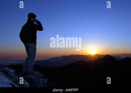 Mann mit Handy bei Sonnenaufgang auf einen Berg oben, Breitenstein Berg, bayerischen Vorbergen, Wendelstein-Gruppe, Oberbayern Stockfoto