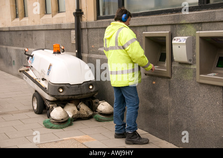 Gemeindeverwaltung Straßenkehrer nehmen Geld aus Geldautomaten Geldautomaten mit seinem Staubsauger an seiner Seite Stockfoto