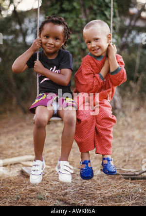 Kleine Jungen und Mädchen sitzen auf zusammen, jeweils eine Seite der Schaukel Seil schwingen, lächelnd in die Kamera, in voller Länge. Stockfoto