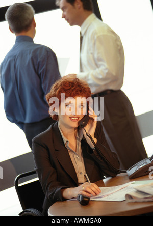 Geschäftsfrau, sitzend, mit Telefon, zwei Geschäftsleute im Hintergrund Stockfoto