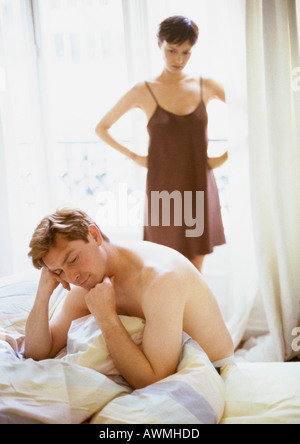 Mann sitzt auf dem Bett, Frau stehend mit Händen auf den Hüften im Hintergrund Stockfoto