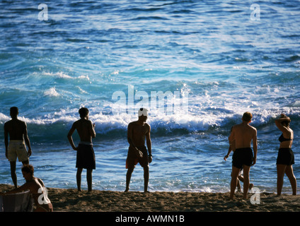 Silhouetten von Menschen stehen am Strand am Rande des Meeres, Wellen im Hintergrund Stockfoto