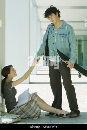 Auf dem Campus schüttelt Studentin am Boden sitzend Hände mit männlichen Studenten, stehend mit skateboard Stockfoto