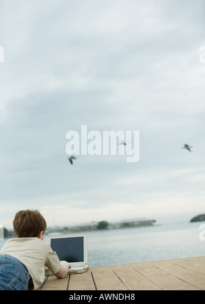 Kind am dock mit Laptop, Vögel fliegen über Wasser im Hintergrund Stockfoto