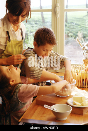 Zwei Kinder und Mutter, die Kinder mit den Händen in die Schüssel, Mutter in Schürze lächelnd in Küche, Kochen Stockfoto