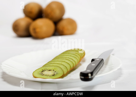 Einzelne geschnittene Kiwi auf einem Teller mit Messer, mehr Kiwis gestapelt auf der Rückseite Stockfoto