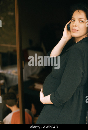 Schwangere Frau stehend, mit Handy, Kind sitzt am Boden im Hintergrund Stockfoto