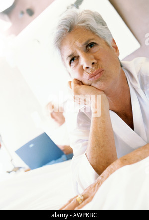 Ältere Frau mit der Hand unter Kinn, Stirn, Mann mit Laptop im Hintergrund Stockfoto