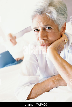 Ältere Frau auf dem Bett mit der Hand unter Kinn, Mann mit Laptop im Hintergrund Stockfoto