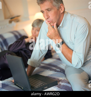 Reifer Mann mit Laptop-Computer auf Bett, Frau im Hintergrund Stockfoto