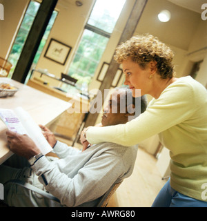 Mann sitzt am Tisch, Frau mit Arme um ihn, Seitenansicht Stockfoto