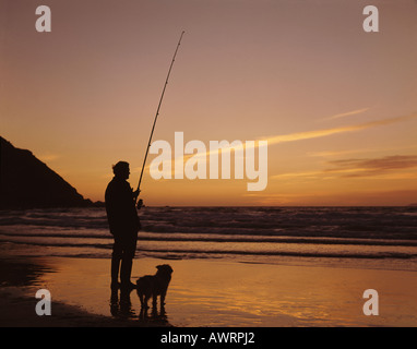 Fliegen Sie Fischer mit der Rute in der hand und Hund an seiner Seite am Rand des Wassers in der Schiffsladung Bay bei Sonnenuntergang in der Nähe von Hartland Point Devon UK Stockfoto