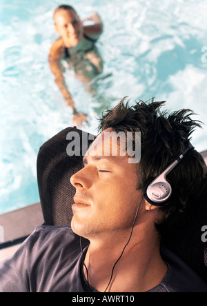 Mann mit Kopfhörern, Frau im Schwimmbad im Hintergrund, erhöhte Ansicht Stockfoto