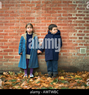 Jungen und Mädchen Hand in Hand und Grimassen vor Mauer, in voller Länge Stockfoto