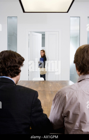Zwei Männer sitzen an einem Tisch und einer Frau, die einen Konferenzraum betreten Stockfoto