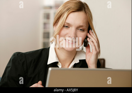 Junge blonde Frau sitzt vor Laptop, Lächeln, als sie am Telefon spricht Stockfoto