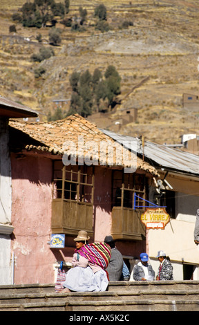 Puno, Peru. Ländliche Quechua-Frau in traditioneller Kleidung sitzen auf Stufen vor einem kleinen Restaurant mit traditionellen Gebäude und Berg... Stockfoto