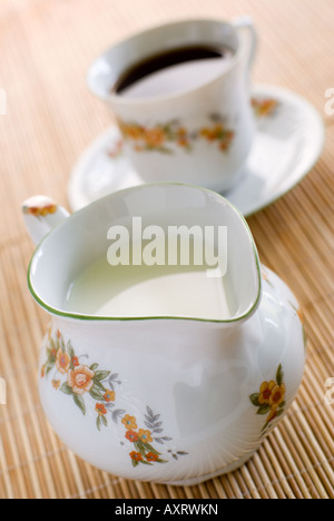 Milch in floralem Muster Porzellan Milchkännchen auf der hölzernen Matte - auf der Vorderseite und die Tasse Kaffee hinter. Flachen DOF. Stockfoto