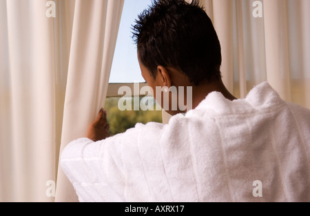 Die junge jamaikanische Frau im Alter von 18 bis 20 Jahren blickt hinter einem Vorhang hervor und blickt nachdenklich durch ein Fenster. Fotografiert in den USA. Stockfoto