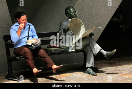 Eine Frau sitzt ihr Mittagessen neben Statue eines Mannes Lesen einer Zeitung in Australien Platz Sydney Australia Stockfoto
