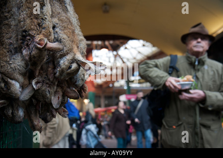 Hasen aufgereiht zum Verkauf in London Borough Market, während ein Mann mit einem Kuchen im Hintergrund vorbeigeht Stockfoto