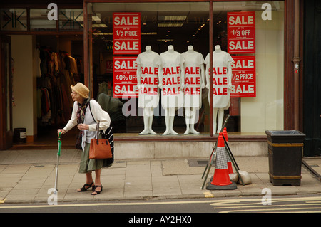 Eine ältere Frau, die mit einem Stock auf dem Bürgersteig an einem Schaufenster mit Verkaufsschildern und Schaufensterpuppen in der High Street, Oxford, vorbeigeht. Stockfoto