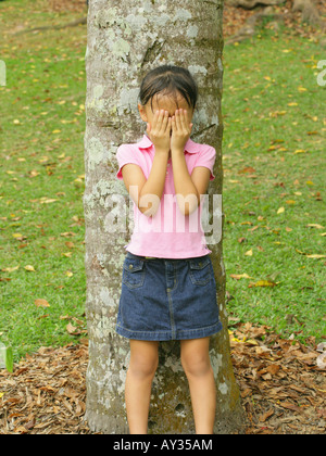 Mädchen steht vor einem Baumstamm und bedeckte ihr Gesicht mit den Händen Stockfoto