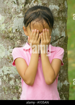 Nahaufnahme eines Mädchens steht man vor einem Baumstamm und bedeckte ihr Gesicht mit den Händen Stockfoto