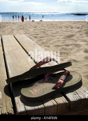 EIN PAAR FLIP-FLOPS SANDALEN AUF EINEM HÖLZERNEN STEG MIT EINEM SONNIGEN STRAND. Stockfoto