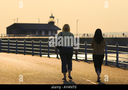 Zwei Frauen zu Fuß auf der Promenade am Morecambe an einem warmen Abend mit vielen fliegenden Insekten mit dem Stein Steg über Stockfoto