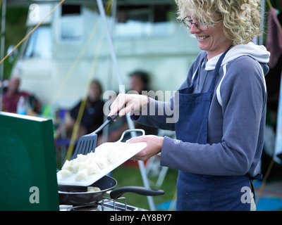 Frau, die Zubereitung von Speisen in einem externen Lager Ambiente mit Gas gefeuert Ofen bereitet Essen mit Zelten und Wohnwagen im Hintergrund lachen Stockfoto