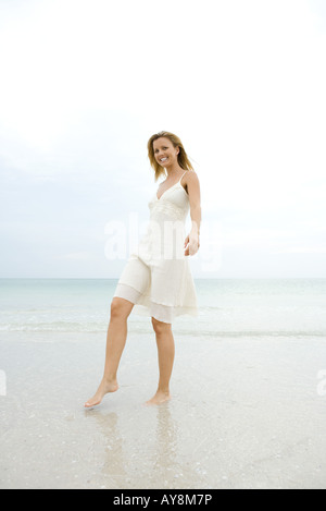 Junge Frau im Sommerkleid zu Fuß auf nassen Sand am Strand, lächelnd in die Kamera, in voller Länge Stockfoto