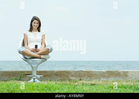 Teenager-Mädchen sitzen auf Stuhl am Meer hält Handy, lächelt in die Kamera Stockfoto