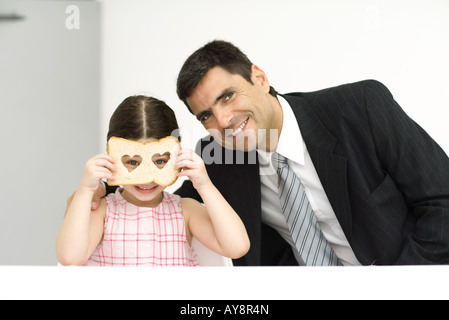 Vater und Tochter sitzen nebeneinander, Mädchen auf der Suche durch Scheibe Brot mit herzförmigen Löcher, beide lächelnd in die Kamera Stockfoto