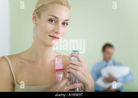 Frau mit Babyflaschen, lächelnd in die Kamera, Mann halten Baby im Hintergrund Stockfoto