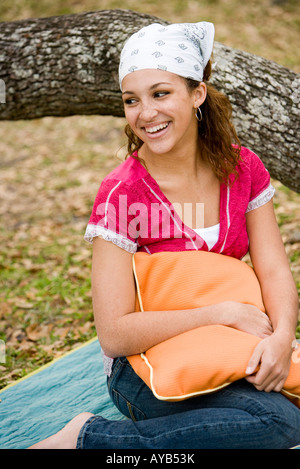 Lächelnde junge Frau mit einem Kissen unter einem Baum sitzt auf einer Decke Stockfoto