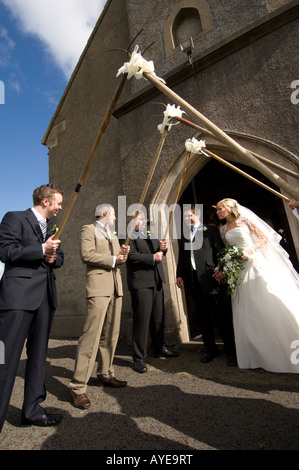 Braut und Bräutigam, die entstehende Kirche durch eine Wache von Ehren af ihre Landwirtschaft Freunde halten Mistgabeln, ländlichen Wales UK Stockfoto