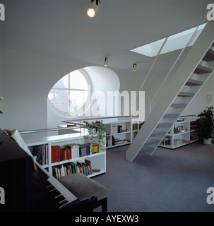 Rundes Fenster über dem niedrigen Bücherregale im modernen weißen Loft Conversion Wohnzimmer mit Klavier und Treppe Stockfoto