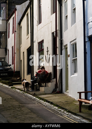 Eine Frau mit einem Hund sitzt in der Sonne Lesen einer Sonntagszeitung außerhalb einer Hütte in Staithes North Yorkshire UK Stockfoto